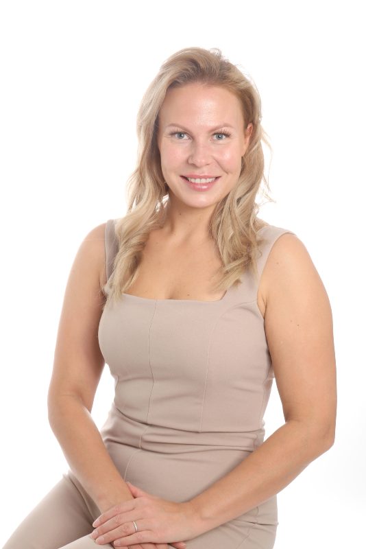 Puolikuva nainen katsoo kameraan, istuu penkillä, kädet sylissä. Iloinen ilme kasvoilla